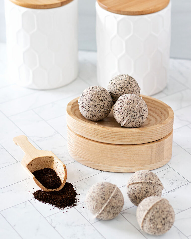 Six coffee bath bombs on a while bathroom countertop with a wooden scoop of ground coffee beans in front of white bath containers.