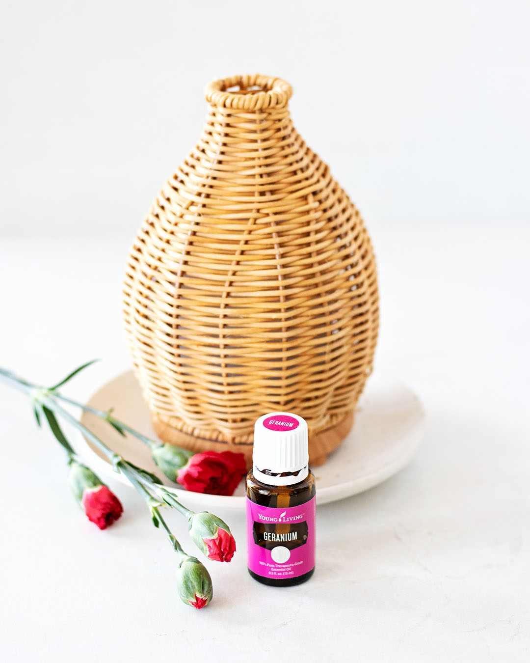 pink bottle of geranium essential oil in front of natural wicker diffuser next to pink red flower stems